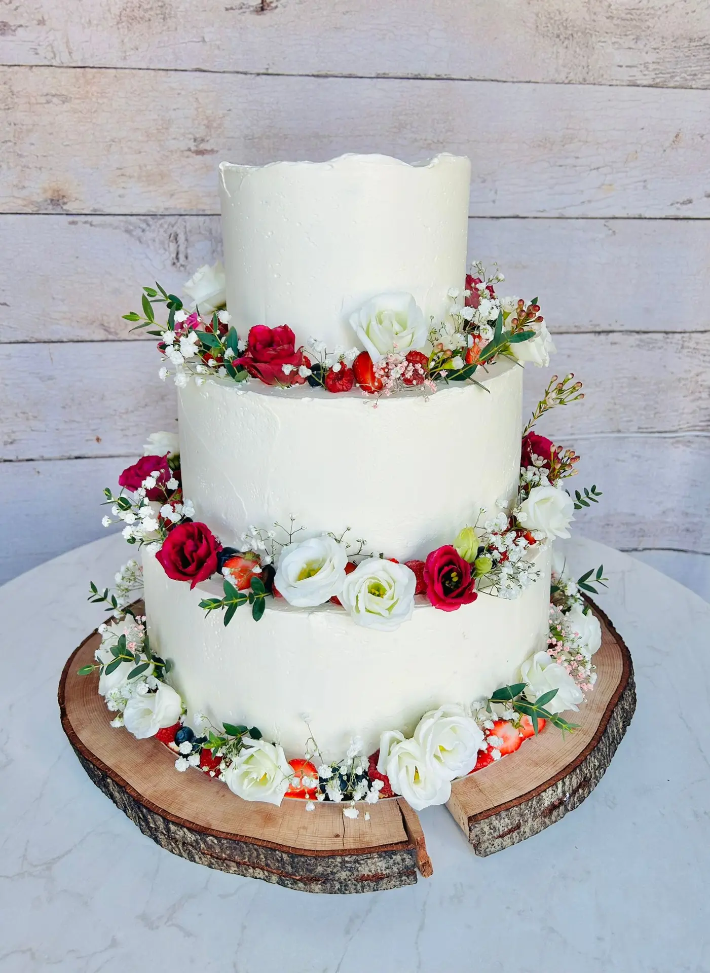 Dreistöckige Hochzeitstorte mit roten und weißen Rosen, Beeren und Schleierkraut auf Holzscheibe